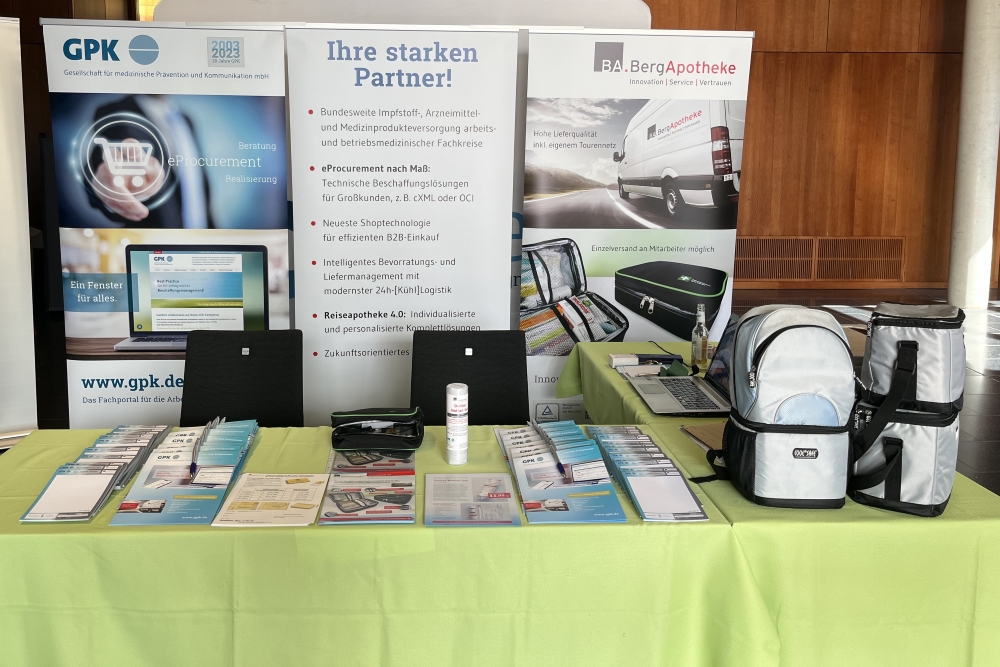 Messestand der GPK und BA.BergApotheke mit Banner 'Ihre starken Partner!' und Informationen zu eProcurement, Reiseapotheke 2.0 und Logistiklösungen, Laptop auf schwarzem Tisch, verschiedene medizinische Produkte auf grünem Tisch.